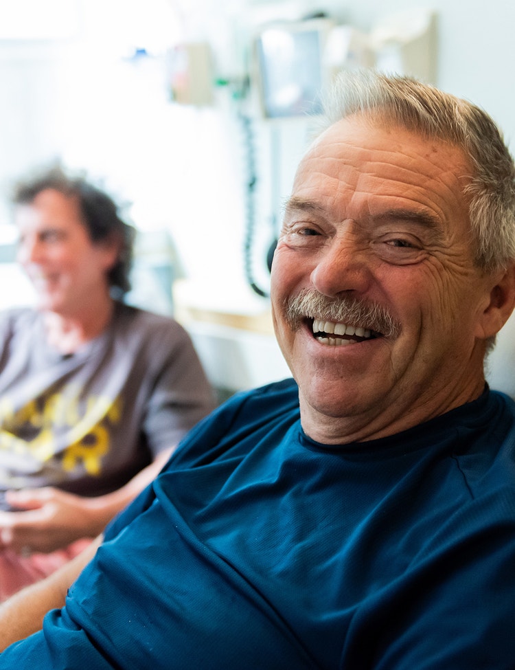 An older man smiles while seated in a clinical setting. Another man sits slightly out of focus in the background.